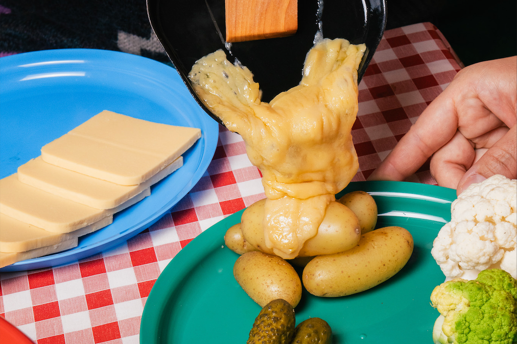 Person using a wooden spatula to pour melted vegan raclette on boiled potatoes. on a checkered tablecloth with plates of cheese, pickles, and vegetables.